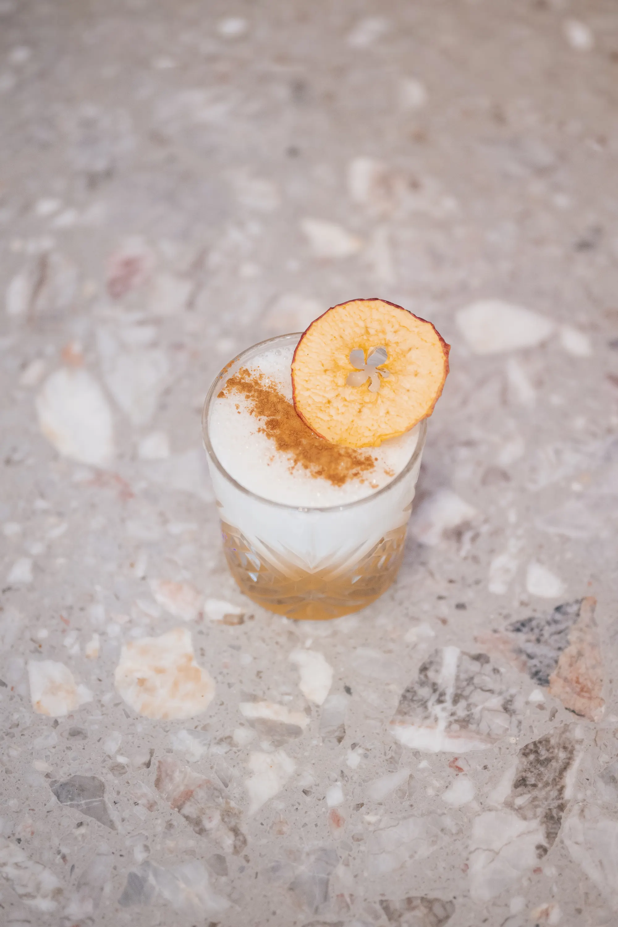Pumpkin Sour sur un comptoir en terrazzo, mousse saupoudrée de cannelle et chips de pomme